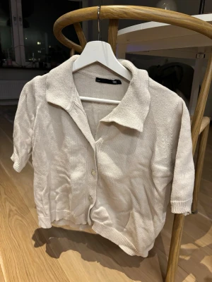 Beige stickad kofta med krage - Säljer en beige stickad kofta från 157 med korta ärmar och klassisk krage. Koftan har knappar framtill och ribbade muddar vid ärmslut och nederkant🐆