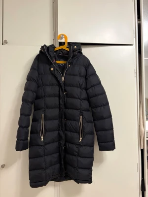 Marinblå lång vinterjacka med huva - Säljer en svart lång dunjacka/vinterjacka med guldiga dragkedjor och knappar. Jackan har huva, två fickor med dragkedja
