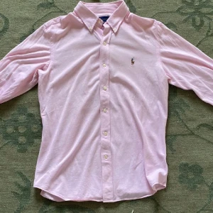 Rosa skjorta från Ralph Lauren - Snygg rosa skjorta från Ralph Lauren i slim fit-modell. Klassisk oxfordstil med knappar hela vägen och långärmad design. Skjortan har broderad logga på bröstet och är gjord i mjuk bomull. Perfekt för dig som gillar stilrena och fräscha plagg.