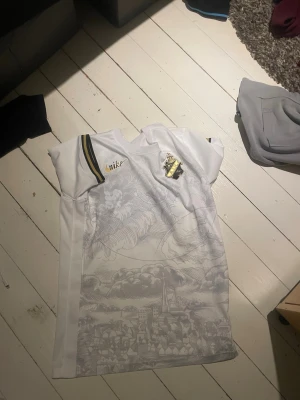 Vit AIK Nike matchtröja - Säljer en vit AIK matchtröja från Nike med snyggt stadsmotiv i grått på framsidan. Tröjan har AIK:s klubbmärke på bröstet och gulsvarta detaljer på ärmarna. Perfekt för dig som vill stötta laget med stil.