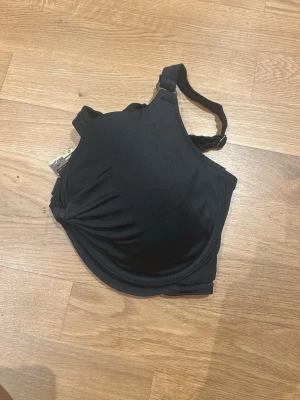 Svart bikiniöverdel från XINT 95D - Svart bikiniöverdel från XINT i storlek 95D. Modellen har formpressade kupor och justerbara axelband. Materialet är mjukt och stretchigt, med en metallspänne som knäppe bak. Perfekt för dig som vill ha stöd och snygg passform på stranden.