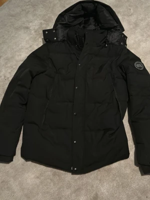 Svart dunjacka från Canada Goose - Svart dunjacka från Canada Goose med huva och snygga detaljer som tryckknappar och dragkedja. Jackan har ett stilrent märke på ärmen och praktiska fickor med dragkedja. Perfekt för kalla dagar och har en clean, modern look.