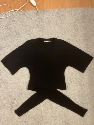 Svart croppad t-shirt med knyt från 157 - Svart croppad t-shirt från 157 med korta ärmar och rund hals. Baksidan har en snygg knyt-detalj som ger extra stilpoäng. Perfekt för dig som gillar enkel men trendig design. Mjuk och skön bomullskvalitet.