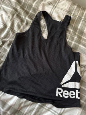 Svart Reebok Speedwick träningslinne - Svart träningslinne från Reebok i storlek L med stort vitt Reebok-logotryck på framsidan. Tillverkat i lätt och ventilerande meshmaterial med Speedwick-teknologi som transporterar bort fukt. Perfekt för gymmet eller löpning.