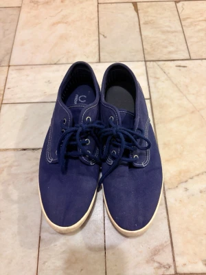 Marinblå sneakers från Fred Perry - Säljer ett par marinblå sneakers från Fred Perry med vita kontrastsömmar och snörning. Skorna har rund tå och vit platt sula. Tillverkade i textilmaterial som ger en avslappnad och stilren look.