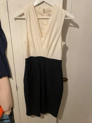 Vit och svart ärmlös klänning  - Snygg ärmlös klänning från H&M i storlek 36. Överdelen är vit med omlott och v-ringad design, medan nederdelen är svart med fickor och veck. Klänningen har dragkedja bak och är tillverkad i ett mjukt syntetmaterial. Klänningen har några små fläckar på framsidan, men dem går säkert bort i tvätten.