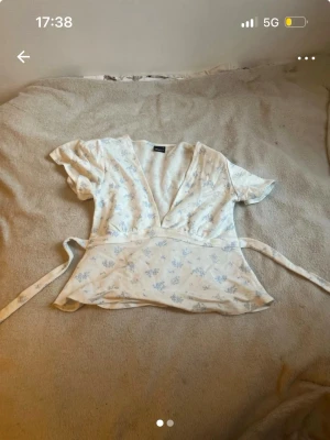 Blommig omlottblus med knytband - Säljer en söt omlottblus från Gina Tricot i ljusbeige med ljusblått blommönster. Blusen har korta ärmar, v-ringning och knytband i midjan för en snygg passform. Perfekt för vår och sommar!