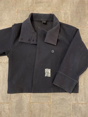 Marinblå kappa - Stilren marinblå kappa från Gina Tricot i storlek S. Jackan har bred krage, dubbelknäppning med knappar. En knapp saknas men extra knappar kommer med i köpet som kan sys fast.