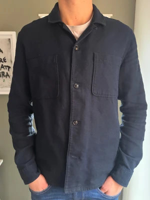 Overshirt | Navy Blue - Snygg mörkblå overshirt från Dressmann med två stora bröstfickor och svarta stilrena knappar.   Perfekt att slänga på över en t-shirt, eller över skjortan för att höja outfiten rejält. Köptes i våras, sen dess använd till och från, grymt skick. Priset är ej hugget i sten.  👕Passform: Storlek M menpassar också L, killen på bilden är 187 cm. 📐Storlek:  M ✅ Skick: 9/10 🧵Material: 100% bomull 🎨 Färg: Navy Blue  ‼️ Skickas samma dag/dagen efter köp‼️ 