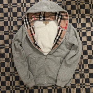Grå Burberry hoodie med dragkedja - Säljer en grå hoodie från Burberry med klassiskt rutigt foder i huvan. Tröjan har dragkedja framtill, snörning i huvan och två fickor. Perfekt för dig som gillar stilren design med en touch av lyx.
