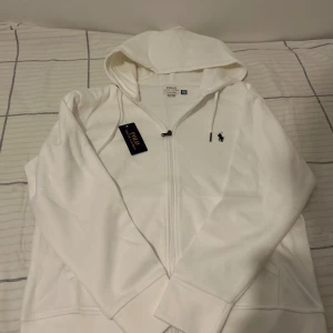 Vit hoodie från Polo Ralph Lauren - Vit hoodie från Polo Ralph Lauren med dragkedja och huva. Klassisk design med svart broderad logga på bröstet, snörning i huvan och fickor framtill. Tillverkad i mjuk bomull, perfekt för chill dagar eller när du vill ha en clean look.