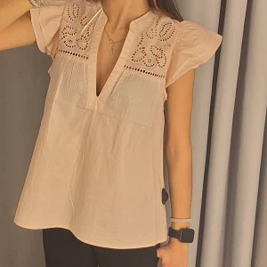 Rosa blus med broderi och volangärm - Supersöt beige blus med broderade detaljer upptill och volangärmar. Blusen har v-ringning och är tillverkad i mjuk bomull.