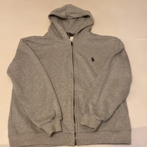Grå hoodie från Polo Ralph Lauren - Grå hoodie från Polo Ralph Lauren med dragkedja framtill och klassisk huva. Köpte den helt ny men var liten för mig
