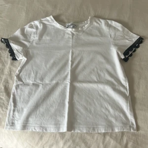 Vit t-shirt med svarta spetsdetaljer Zara - Vit t-shirt från Zara i bomull med korta ärmar och snygga svarta spetsdetaljer längst ut på ärmarna.