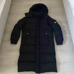 Lång Moncler jacka  - Säljer en svart lång dunjacka med huva och dragkedja framtill. Jackan har två snedställda fickor med dragkedja och Moncler-logga på ärmen. Perfekt för kalla dagar när du vill hålla dig varm och snygg.