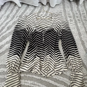 Randig blus från NA-KD i svart och vitt - Säljer en snygg blus från NA-KD i storlek 38. Den har svartvita ränder, v-ringning och knappar framtill. Blusen har långa ärmar med breda manschetter och är tillverkad i ett mjukt, glansigt material som känns lyxigt.