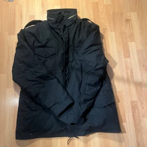 Svart parkajacka från Bandit Vintage XL - Svart parkajacka från Bandit Vintage Clothing i storlek XL. Jackan har hög krage med dragkedja, flera stora fickor framtill och axelremmar. Tillverkad i slitstark bomull med fodrad insida för extra komfort. Perfekt för dig som gillar en klassisk och stilren look. Nypris 1500