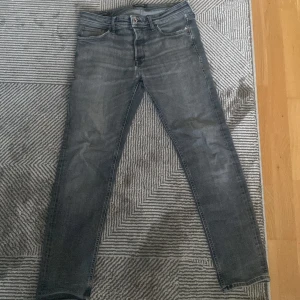 Grå skinny jeans från Jack & Jones - Snygga grå jeans från Jack & Jones med smal passform och klassisk femficksdesign. Jeansen har en något tvättad look och är tillverkade i mjukt denimtyg. Perfekta för dig som gillar en modern och stilren vibe. Jeansen har några små hål vid skrevet som bör lagas