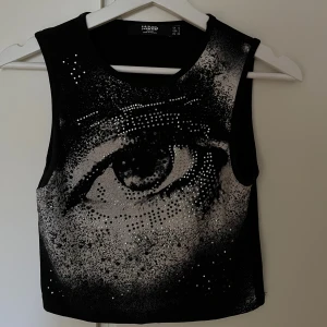 Svart croppad topp med ögonmotiv Jaded - Svart croppad topp från Jaded London med ett stort ögonmotiv framtill, dekorerad med glittriga stenar. Kan mötas upp på Södermalm!