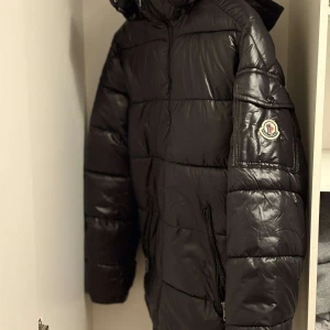 Svart Moncler pufferjacka M - Säljer en svart pufferjacka från Moncler i storlek M. Jackan har glansig finish, klassisk Moncler-logga på ärmen, dragkedja framtill och två fickor med dragkedja. Fodret är i svart och har en unik serietidningsdetalj på insidan. Perfekt för kalla dagar.