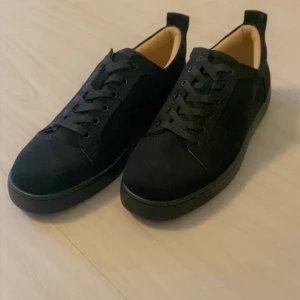Svarta sneakers från Christian Louboutin - Snygga svarta sneakers från Christian Louboutin i mocka med klassisk snörning och rund tå. Skorna har ikoniska röda sulor med logotyp. Insidan är ljusbeige och materialet känns lyxigt och stilrent.