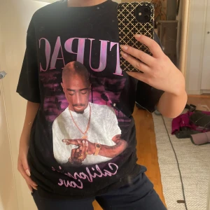 Svart Tupac t-shirt med tryck - Säljer en svart t-shirt med stort Tupac-tryck framtill och lila text. Oversized passform och korta ärmar. Perfekt statement-plagg för dig som gillar hiphop och streetstyle.