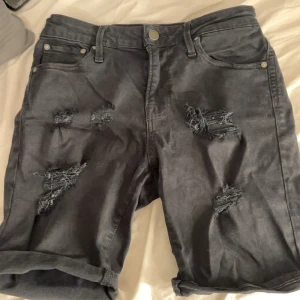 Svarta jeansshorts Jack & Jones - Svarta jeansshorts från Jack & Jones med snygga slitningar och upprullade benslut. Klassisk femficksmodell med knapp och dragkedja. Regular fit och cool streetkänsla, perfekta för sommaren.