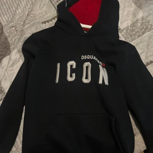 Svart Dsquared2 ICON hoodie med röd huva - Svart hoodie från Dsquared2 med stor vit 'ICON'-text och broderad logga på bröstet. Röd insida på huvan och snygga Dsquared2-detaljer på ärmen och ryggen. Klassisk känguruficka och lång ärm. Perfekt för dig som gillar streetwear vibes.