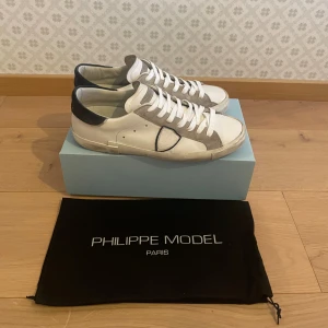 Philippe Model vita sneakers med grå detaljer - Snygga vita Philippe Model sneakers med grå mockadetaljer och mörkblå häl. Klassisk låg modell med snörning och rund tå. Skorna är i skinn och mocka, och levereras med originalpåse och låda. Perfekta för en stilren och trendig look. Kan tänka mig att gå ner i pris. Är i mycket bra skick, storlek 44 hör av dig om du undrar något mvh Levi
