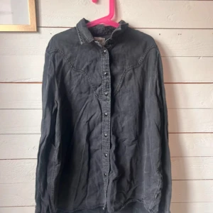 Svart jeansskjorta från Lee - Säljer en svart jeansskjorta från Lee med relaxed fit. Skjortan har klassisk krage, tryckknappar framtill och långa ärmar. Snygga sömdetaljer över bröstet ger en cool westernkänsla. Perfekt att styla lager på lager eller bära som den är.