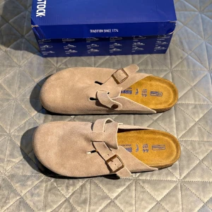 Birkenstock Boston grey suede mocka sandaler - Birkenstock Boston sandaler i grey suede mocka med justerbart spänne i metall. Skorna har en mjuk fotbädd och en platt sula med mönstrad undersida för extra grepp. Perfekt för chill dagar och enkel att slänga på sig. Klassisk rund tå och stilren design.