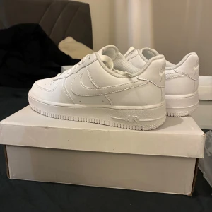 Nike Air Force 1 helvita sneakers - Klassiska Nike Air Force 1 sneakers i helvitt skinn med låg profil och snygg, clean design. Ikonisk swoosh på sidorna, perforerad tå och platt sula. Perfekta för dig som vill ha en tidlös och fräsch look till vardagsoutfiten.