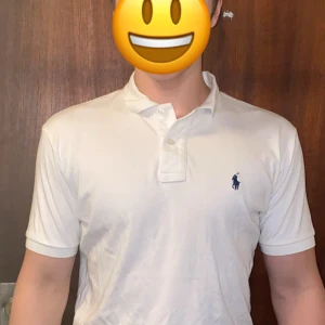 Vit pikétröja från Ralph Lauren - Klassisk vit pikétröja från Ralph Lauren med korta ärmar och blå broderad logga på bröstet. Tröjan har krage och knäppning med två knappar framtill. Perfekt för en clean och stilren look. Modellen på bilden är 183cm lång och väger ungefär 80kg.
