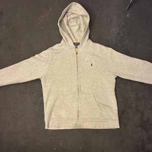 Grå hoodie från Polo Ralph Lauren Junior XL - Klassisk grå hoodie från Polo Ralph Lauren i storlek junior XL. Tröjan har dragkedja, huva och två fickor framtill. Broderad marinblå logga på bröstet. Perfekt för chill dagar och enkel att matcha med allt. Tillverkad i mjuk bomullsmix.