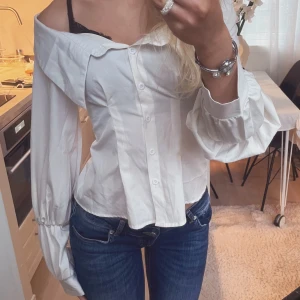 Vit offshoulder skjorta med puffärm - Säljer en vit skjorta med offshoulder-design och puffiga långärmar. Skjortan har knappar framtill och en snygg, figurnära passform. 