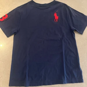 Mörkblå t-shirt Polo Ralph Lauren - Mörkblå t-shirt från Polo Ralph Lauren med röd broderad logga på bröstet och en röd siffra 3 på ärmen. Klassisk rund hals och normal passform. Tillverkad i mjuk bomull, perfekt för en clean och stilren look.