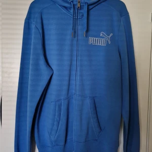 Blå hoodie från Puma XL - Blå hoodie från Puma i storlek XL med dragkedja och huva. Tröjan har fickor framtill och Puma-logga på bröstet. Perfekt för chill dagar eller när du vill ha en sportig look. Tillverkad i mjukt material för extra komfort.