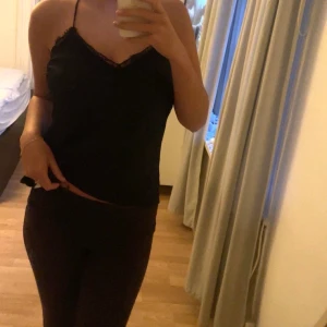 Svart satin topp med spets - Svart topp i satin med tunna axelband och v-ringning dekorerad med spets. Modellen har smala band,  korsade band i ryggen och har en enkel, stilren design som passar perfekt för en trendig look. Tunn och lätt i materialet, vilket gör den skön att bära under varma dagar.