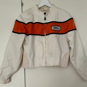 Harley-Davidson racingjacka vit/orange - Snygg racinginspirerad jacka från Harley-Davidson i vit med bred orange panel och svarta detaljer. Jackan har dragkedja framtill, hög krage med knapp och broderad logga på bröstet och ryggen. Materialet är slitstark bomull och polyester. Jackan har slitage i form av fläckar (se bilder) men går att rengöra eller kemtvätta. I övrigt är den i bra skick. Har strl S men har burit den oversized! 