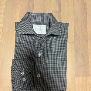 Rutig skjorta från Oscar Jacobson - Snygg svartvit rutig skjorta från Oscar Jacobson i 100% bomull. Skjortan har lång ärm, knappar framtill och vid manschetten, och är tillverkad i mjuk bomull som känns skön mot huden.  Observera att det finns ett litet hål som inte syntes vid användning.  Hör gärna av dig vid frågor, mått eller om du vill ha fler bilder.