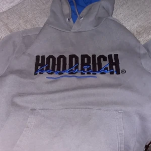 Grå HOODRICH hoodie med blå detaljer - Säljer en grå hoodie från HOODRICH i storlek S med svart och blå broderad logga framtill och tryck på ryggen. Hoodien har blått innerfoder i huvan och klassisk känguruficka. Perfekt för dig som gillar streetwear och vill sticka ut. ( finns en liten defekt som syns på bild)