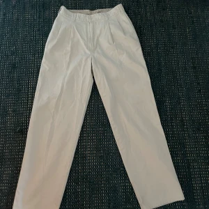 Beiga chinos från Dockers - Snygga beiga chinos från Dockers med klassisk rak passform och hög midja. Byxorna har en sönder dragkedja i gylfen och missar en knapp, pressveck framtill och bakfickor med knapp. Perfekta för en clean och stilren look.