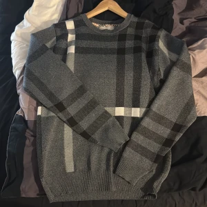 Grå rutig tröja från Burberry - Säljer en snygg grå tröja från Burberry med klassiskt rutigt mönster i svart, vitt och grått. Tröjan har rund halsringning, långa ärmar och ribbade muddar. Perfekt för dig som gillar stilrena och tidlösa plagg.