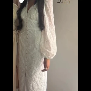 Vit långklänning med spets och puffärm - Supersnygg vit långklänning från NA-KD med spetsdetaljer och puffiga långärmar. Klänningen har v-ringning, markerad midja och knappar framtill samt en slits. Perfekt för dig som vill ha en romantisk och trendig look.