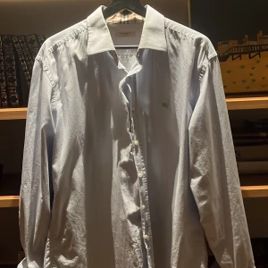 Ljusblå skjorta från Burberry - Snygg ljusblå skjorta från Burberry med klassisk passform. Skjortan har långa ärmar, knappar framtill och ikoniskt rutigt tyg på insidan av kragen. Broderad Burberry-logga på bröstet. Perfekt för dig som gillar stilrena och exklusiva plagg.