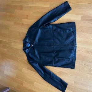 Skinnjacka från Tweed River  - Snygg svart skinnjacka från Tweed River i äkta läder med klassisk krage och dragkedja framtill. Jackan har raka linjer, två fickor och helsvart foder i polyester. Perfekt för dig som vill ha en stilren och tidlös look.