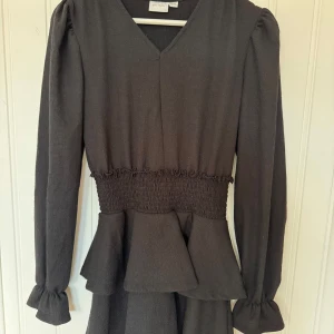 Svart volangklänning från Gina Tricot - Fin svart klänning från Gina Tricot med v-ringning, långa ärmar och markerad midja med smock. Volangdetaljer vid midjan.