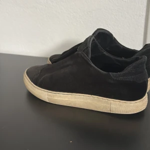 Svarta sneakers från Axel Arigato - Svarta sneakers från Axel Arigato i mocka med vit platt sula. Skorna har en minimalistisk design utan snörning och en diskret logga på plösen. Perfekta för dig som gillar stilrena och bekväma skor med en modern touch. Sänker priset vid snabb affär