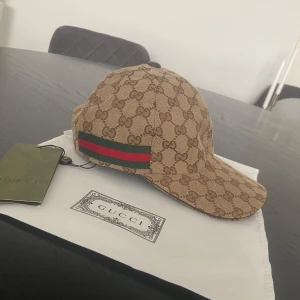 Beige gucci keps - Exklusiv keps från Gucci i beige med klassiskt GG-monogram över hela kepsen. Sidorna har den ikoniska gröna och röda randen. Justerbar rem baktill i svart läder. Tillverkad i bomullscanvas med mörkbrunt innerfoder. Perfekt för dig som vill sticka ut med lyxig streetstyle.