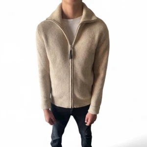 Beige stickad kofta med dragkedja - Säljer en beige stickad kofta med ribbad struktur och hel dragkedja framtill. Koftan har hög krage och långa ärmar, perfekt för lager-på-lager under kyliga dagar. Enkel och clean design som funkar till det mesta. Märket är Arne Clothing. 